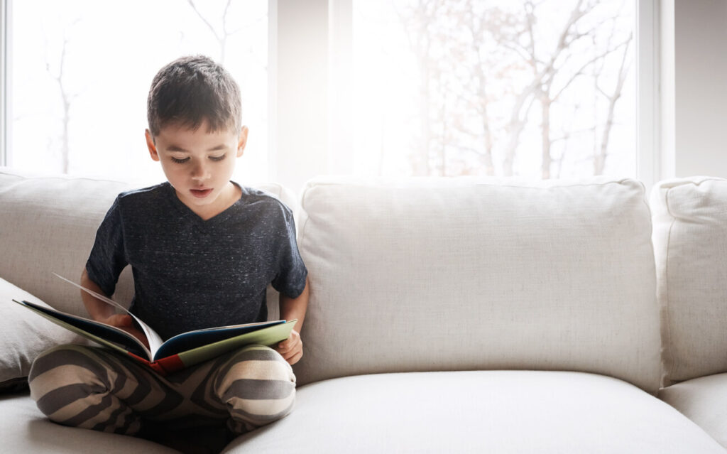 Menino lendo no sofá durante as férias escolares com livros espíritas infantis.